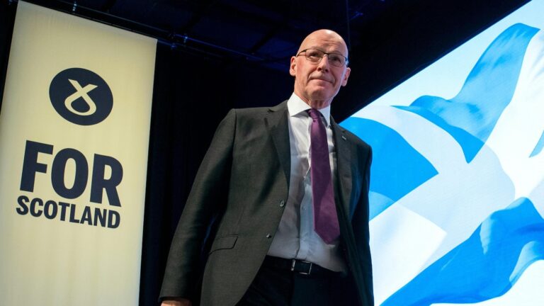 Scotland's First Minister and leader of the SNP, John Swinney MSP, on stage at SNP Conference. He wears a dark suit and purple tie, there is a yellow and black sign with the SNP symbol and "For Scotland" in large letters to his right and an image of Scotland's flag to his left.