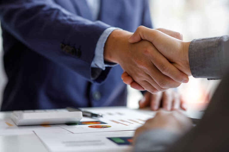 People wearing professional clothes shaking hands. Papers showing graphs and charts can be seen in front of them.