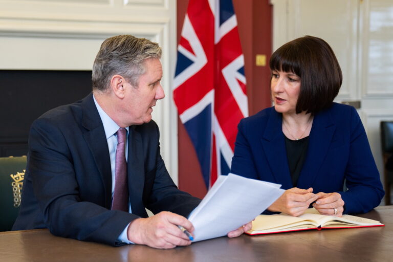 Keir Starmer, Leader of the Labour Party, and Rachel Reeves, Shadow Chancellor of the Exchequer, sit at a polished wood table. Turned slightly to face each other, they are in conversation. Each has papers in front of them and there is a Union Jack flag in the background.