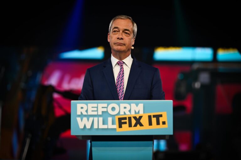 Leader of Reform UK, Nigel Farage, is standing at a podium delivering a speech at the Utilita Arena Birmingham, 28 March 2025. He is dressed in a suit and is addressing an audience.
