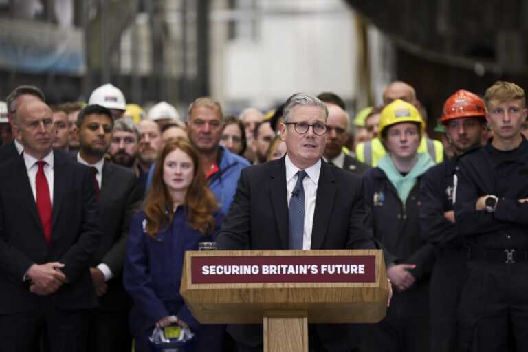 Prime Minister Keir Starmer gives a press conference as he and the Defence Secretary John Healey visits HMS Glasgow, as the government unveils its defence spending plans. The Prime Minister stands confidently at a podium, smiling as he addresses a large crowd of people behind him.