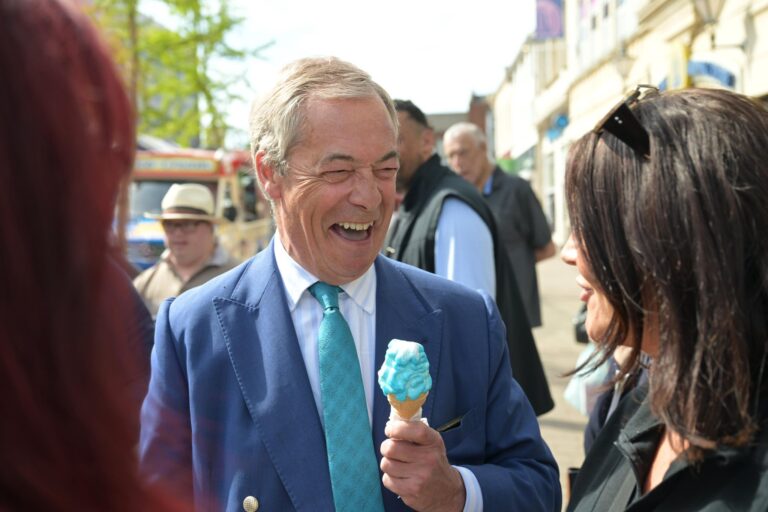 Nigel Farage, Leader of Reform UK, in a blue suit with a turquoise tie, joyfully eating a blue ice cream cone. He is smiling broadly, standing on a sunny street with a crowd in the background. A woman with shoulder-length dark hair, wearing sunglasses, stands in front of him.