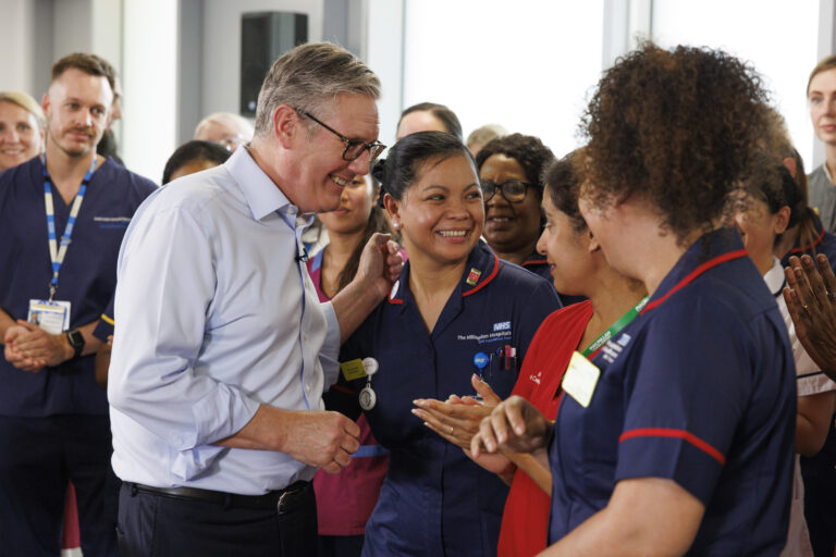 A group of smiling healthcare workers in blue uniforms, gathered around Prime Minister Keir Starmer who is wearing glasses wearing a light blue shirt. The atmosphere is warm and celebratory.