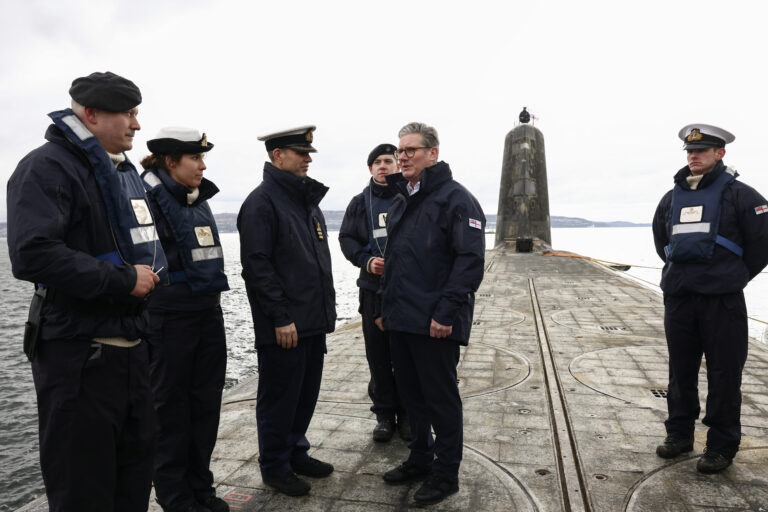 Prime Minister Keir Starmer visits a Vanguard class submarine off the coast of Scotland as it returns home from a period of duty at sea. He is standing on the submarine wearing a dark blue outdoors jacket, members of the armed services stand around him in conversation.