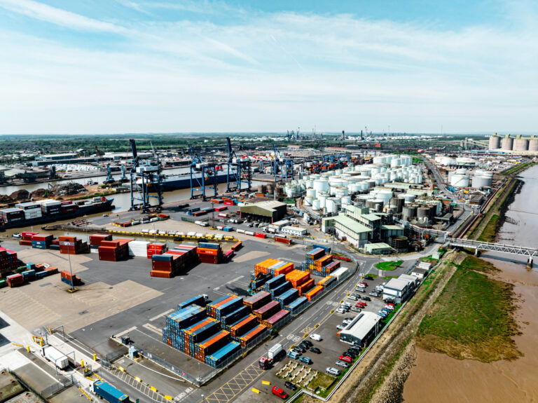 Aerial view of a busy container port with blue gantry cranes, stacked shipping containers, and rail tracks sprawling across the yard.