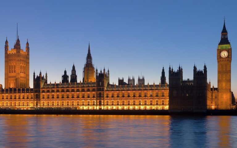 Evening view of the Palace of Westminster and Big Ben along the Thames, illuminated against a blue sky.