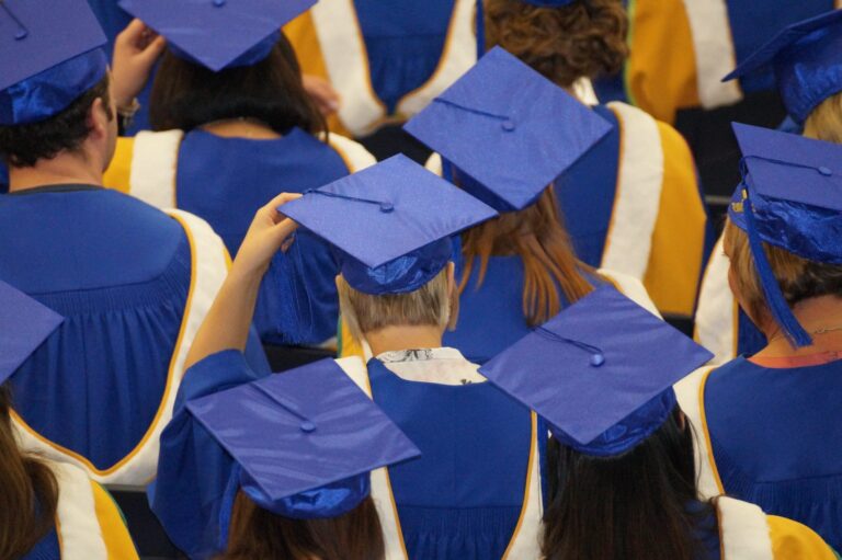 Graduates in blue caps and gowns at a commencement ceremony, seen from behind.
