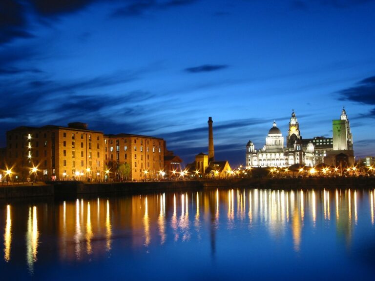 Night view of a illuminated riverfront city skyline at dusk, with brick warehouses on the left and a bright domed building on the right reflecting in the water.